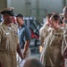 A group of U.S. Navy personnel in tan/khaki service uniforms are gathered in what appears to be a hangar or industrial facility. Two central figures — a senior enlisted male sailor wearing a combination cover and a female sailor — appear to be engaged in a friendly conversation, both smiling. The setting suggests an informal gathering or post-ceremony interaction.
