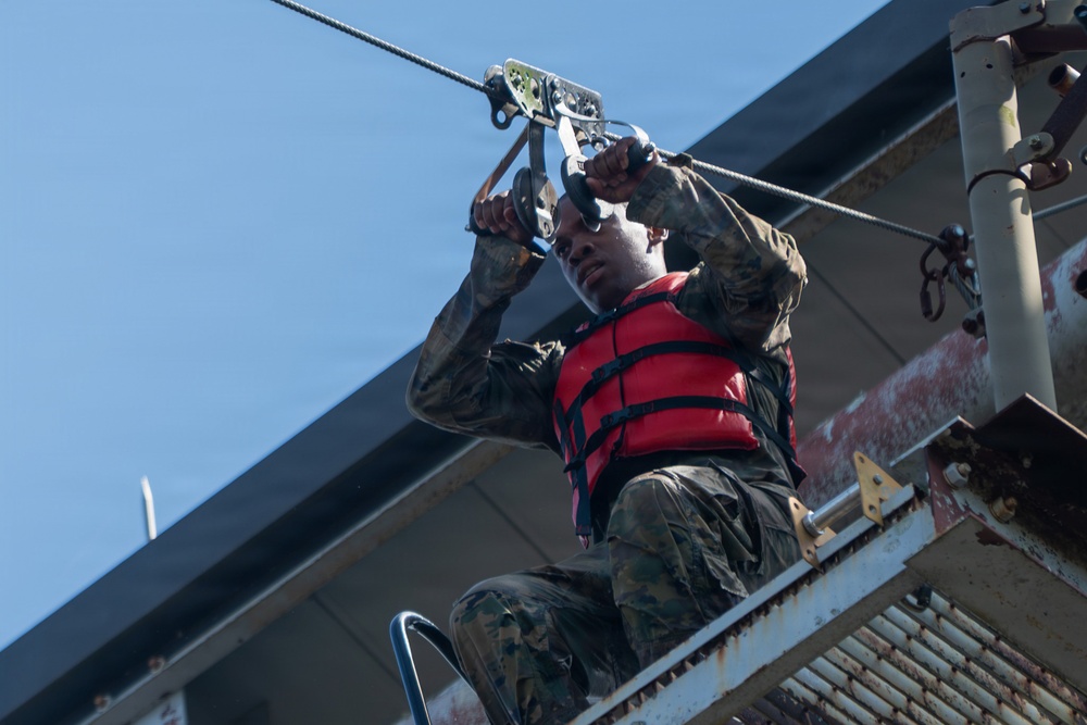 A person wearing military camouflage uniform and a red life vest is preparing to use a zip line or cable traverse system on an elevated metal platform. They are gripping a pulley/trolley device attached to a steel cable overhead. The setting appears to be a military training facility or obstacle course near water.