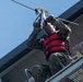 A person wearing military camouflage uniform and a red life vest is preparing to use a zip line or cable traverse system on an elevated metal platform. They are gripping a pulley/trolley device attached to a steel cable overhead. The setting appears to be a military training facility or obstacle course near water.