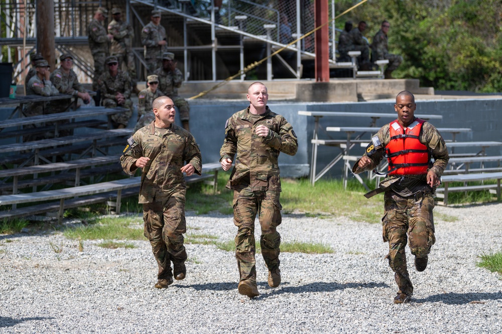 Three soldiers in US Army camouflage uniforms are running across a gravel surface during what appears to be a military training exercise or competition. The soldier on the right is wearing a red life vest and appears wet, suggesting a water-based obstacle or event was recently completed. A group of uniformed military personnel observes from bleachers in the background.