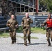 Three soldiers in US Army camouflage uniforms are running across a gravel surface during what appears to be a military training exercise or competition. The soldier on the right is wearing a red life vest and appears wet, suggesting a water-based obstacle or event was recently completed. A group of uniformed military personnel observes from bleachers in the background.