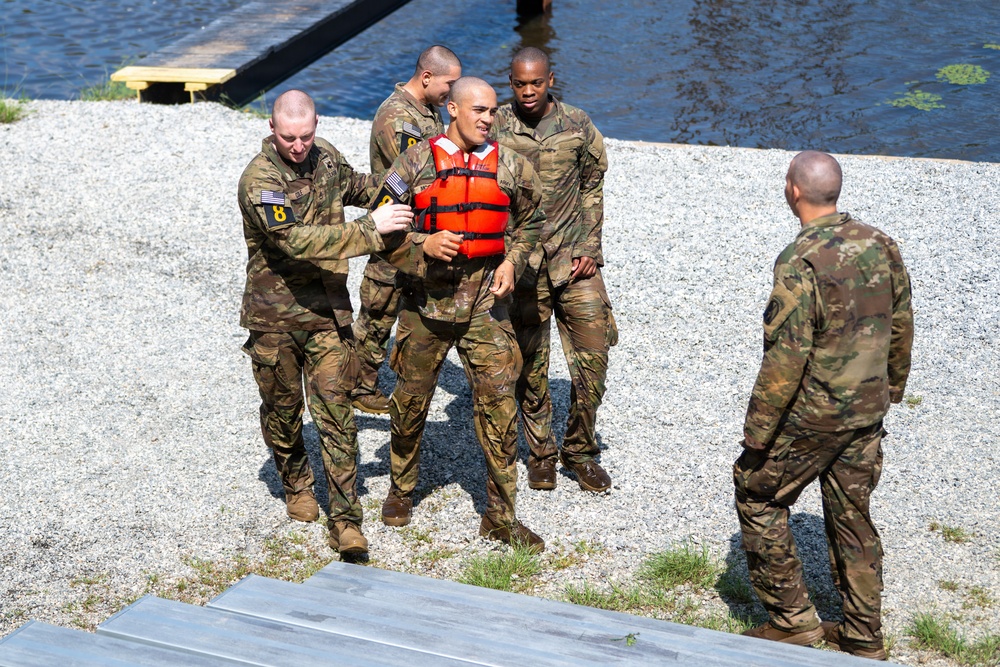 A group of military personnel in camouflage uniforms are assisting a fellow soldier wearing an orange life vest near a body of water on a gravel shoreline. The soldier in the life vest appears to have just emerged from the water and is being supported by two or three comrades. A fifth soldier stands nearby observing the situation.