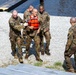 A group of military personnel in camouflage uniforms are assisting a fellow soldier wearing an orange life vest near a body of water on a gravel shoreline. The soldier in the life vest appears to have just emerged from the water and is being supported by two or three comrades. A fifth soldier stands nearby observing the situation.