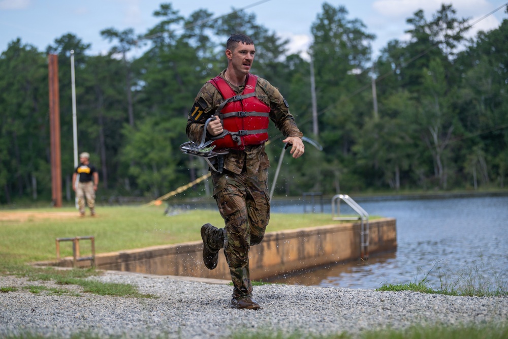 A soldier in wet camouflage uniform and red life vest is running along a gravel path near a water obstacle course or training pond. The individual appears to have just exited the water, as evidenced by dripping water and mud-soaked clothing. This appears to be a military training exercise or competition event.
