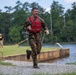 A soldier in wet camouflage uniform and red life vest is running along a gravel path near a water obstacle course or training pond. The individual appears to have just exited the water, as evidenced by dripping water and mud-soaked clothing. This appears to be a military training exercise or competition event.