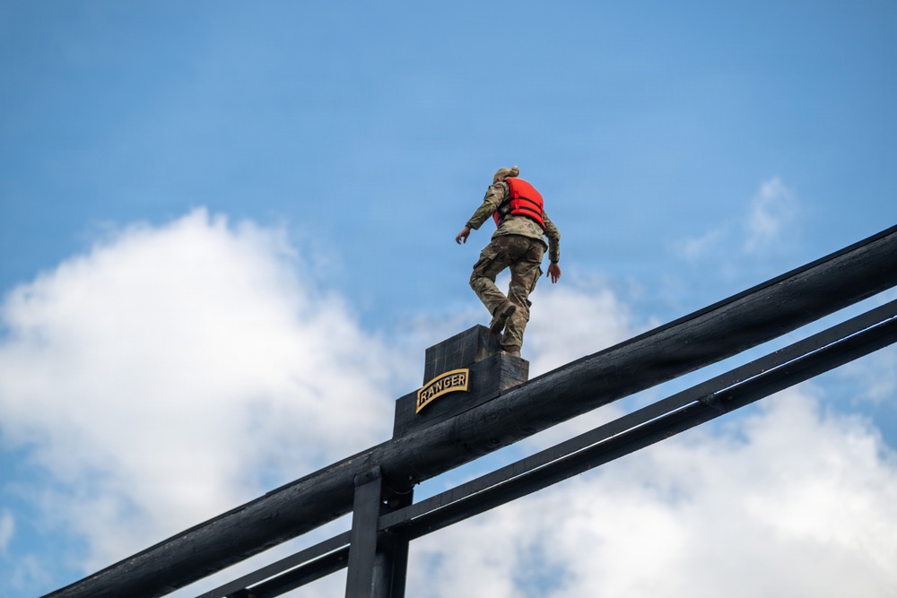 A soldier in camouflage uniform and red life vest stands atop a tall black metal structure marked with a 'RANGER' tab, photographed from a low angle against a partly cloudy blue sky. The image appears to capture a U.S. Army Ranger training exercise, likely involving a water obstacle or high-structure jump. The low-angle perspective emphasizes the height and intensity of the activity.