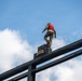 A soldier in camouflage uniform and red life vest stands atop a tall black metal structure marked with a 'RANGER' tab, photographed from a low angle against a partly cloudy blue sky. The image appears to capture a U.S. Army Ranger training exercise, likely involving a water obstacle or high-structure jump. The low-angle perspective emphasizes the height and intensity of the activity.