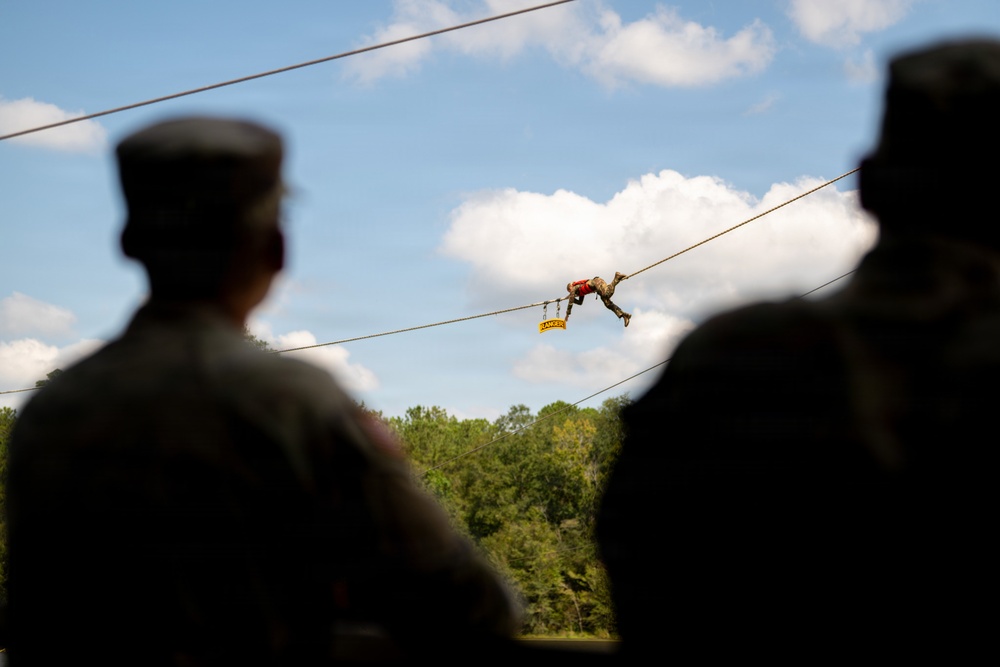 Two silhouetted military personnel in uniform caps observe a soldier traversing a rope/cable line in the background. The active soldier appears to be performing a commando-style rope crossing or zip-line exercise, with a yellow 'RANGER' tab/sign visible hanging from the cable. The scene takes place at what appears to be a military training facility surrounded by trees under a partly cloudy sky.