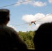Two silhouetted military personnel in uniform caps observe a soldier traversing a rope/cable line in the background. The active soldier appears to be performing a commando-style rope crossing or zip-line exercise, with a yellow 'RANGER' tab/sign visible hanging from the cable. The scene takes place at what appears to be a military training facility surrounded by trees under a partly cloudy sky.
