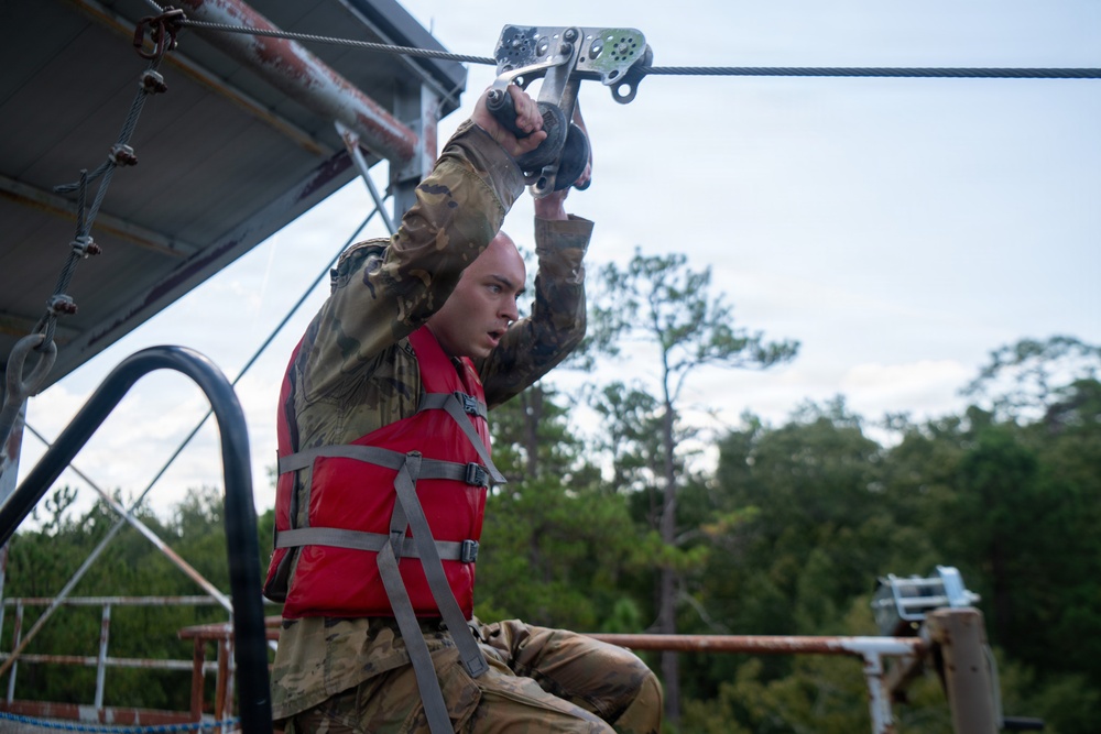 A young male in military camouflage uniform and a red life vest grips a zip-line pulley mechanism overhead, appearing to be at the launch point of a water obstacle course. The setting appears to be a military training facility with a raised platform structure over or near water. Trees and an overcast sky are visible in the background.