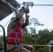 A young male in military camouflage uniform and a red life vest grips a zip-line pulley mechanism overhead, appearing to be at the launch point of a water obstacle course. The setting appears to be a military training facility with a raised platform structure over or near water. Trees and an overcast sky are visible in the background.