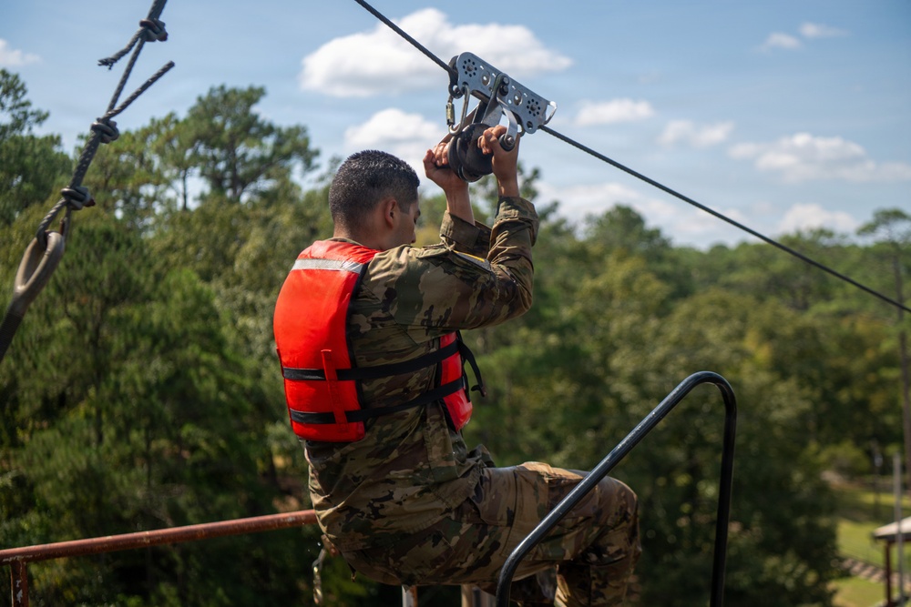 A person wearing military camouflage uniform and an orange life vest is positioned at the top of a zipline platform, gripping a metal trolley/pulley handle attached to the cable. The individual appears to be preparing to launch from an elevated platform surrounded by dense green trees under a partly cloudy sky. The setting suggests a military training exercise involving a water crossing or obstacle course.