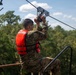 A person wearing military camouflage uniform and an orange life vest is positioned at the top of a zipline platform, gripping a metal trolley/pulley handle attached to the cable. The individual appears to be preparing to launch from an elevated platform surrounded by dense green trees under a partly cloudy sky. The setting suggests a military training exercise involving a water crossing or obstacle course.