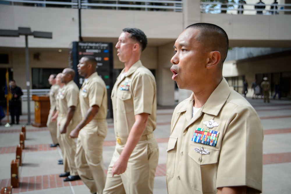A group of U.S. Navy personnel in tan/khaki service uniforms are standing in formation at what appears to be an outdoor military ceremony. The foreground sailor displays multiple service ribbons, warfare qualification badges, and other decorations on his uniform. The setting appears to be a plaza or courtyard of a military or government facility.