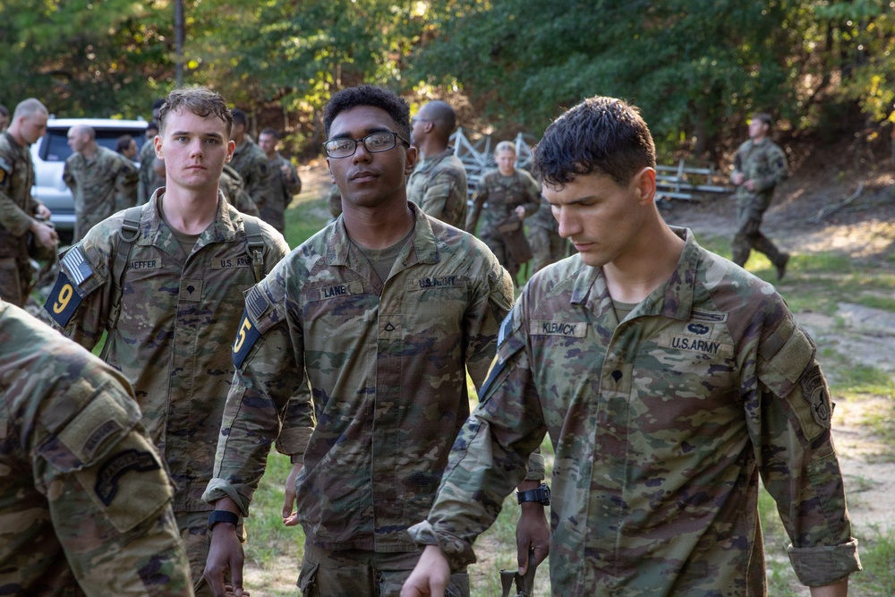 A group of U.S. Army soldiers in OCP (Operational Camouflage Pattern) uniforms are gathered in an outdoor wooded training area. Three soldiers are prominently visible in the foreground with name tapes reading 'SCHAEFFER', 'LANE', and 'KLEMICK', all wearing U.S. Army insignia. The setting appears to be a field training exercise or military training event with numerous other soldiers visible in the background.