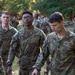 A group of U.S. Army soldiers in OCP (Operational Camouflage Pattern) uniforms are gathered in an outdoor wooded training area. Three soldiers are prominently visible in the foreground with name tapes reading 'SCHAEFFER', 'LANE', and 'KLEMICK', all wearing U.S. Army insignia. The setting appears to be a field training exercise or military training event with numerous other soldiers visible in the background.