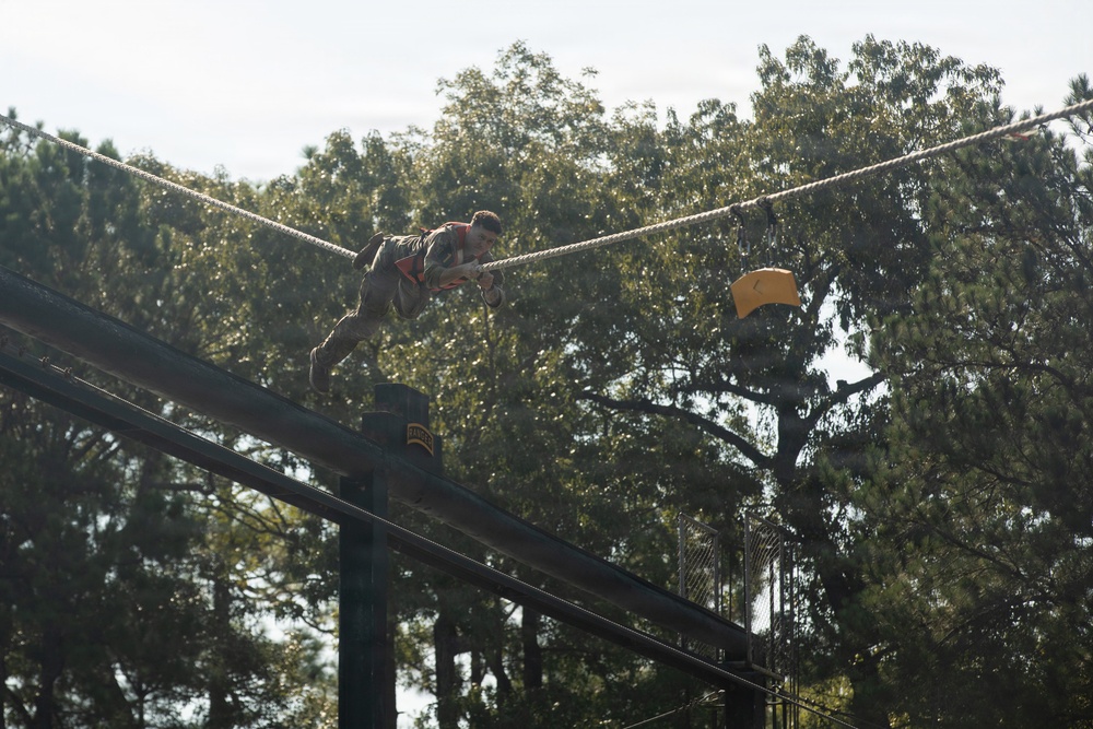 A person is traversing a high rope obstacle course element, gripping a diagonal rope while suspended at elevation above a wooden platform structure. A yellow marker or flag is visible on the right side of the frame. The setting appears to be a military or fitness training facility surrounded by tall trees.