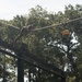 A person is traversing a high rope obstacle course element, gripping a diagonal rope while suspended at elevation above a wooden platform structure. A yellow marker or flag is visible on the right side of the frame. The setting appears to be a military or fitness training facility surrounded by tall trees.