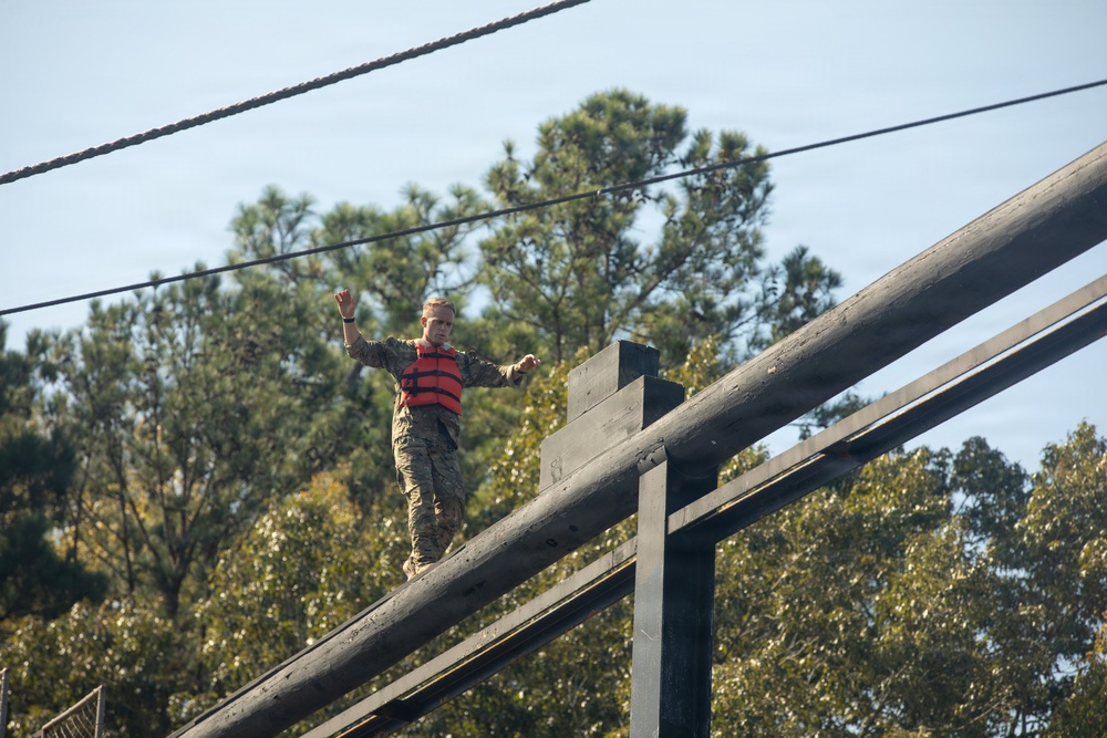 A person in military camouflage uniform and an orange life vest is balancing on a elevated wooden beam or log structure, arms outstretched for balance. The setting appears to be a military obstacle course or training facility, likely near water given the life vest requirement. Trees and a clear sky are visible in the background.