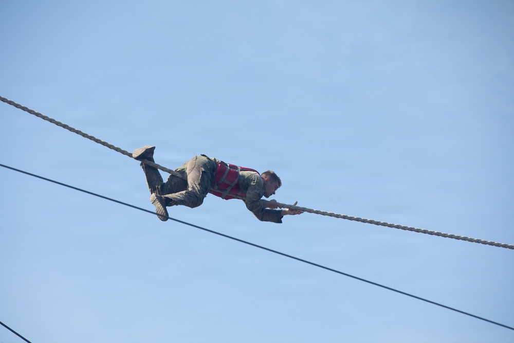 A person is traversing a rope bridge or obstacle course high in the air, suspended between two ropes against a clear blue sky. They are in a horizontal position, gripping an upper rope with their hands while hooking their feet over the same or a parallel rope. The individual is wearing military-style camouflage clothing and a red safety harness vest.
