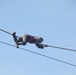 A person is traversing a rope bridge or obstacle course high in the air, suspended between two ropes against a clear blue sky. They are in a horizontal position, gripping an upper rope with their hands while hooking their feet over the same or a parallel rope. The individual is wearing military-style camouflage clothing and a red safety harness vest.