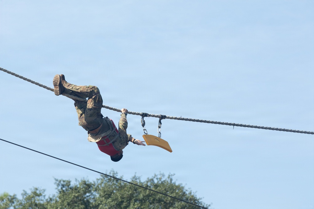 A person in military camouflage uniform and combat boots is hanging upside down from a rope or cable suspended high in the air, appearing to traverse or navigate an aerial obstacle course. They are wearing a red harness and are connected to the rope via carabiners and rigging hardware. A yellow curved object, likely a handle or grip device, hangs nearby on the same rope system.