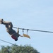 A person in military camouflage uniform and combat boots is hanging upside down from a rope or cable suspended high in the air, appearing to traverse or navigate an aerial obstacle course. They are wearing a red harness and are connected to the rope via carabiners and rigging hardware. A yellow curved object, likely a handle or grip device, hangs nearby on the same rope system.