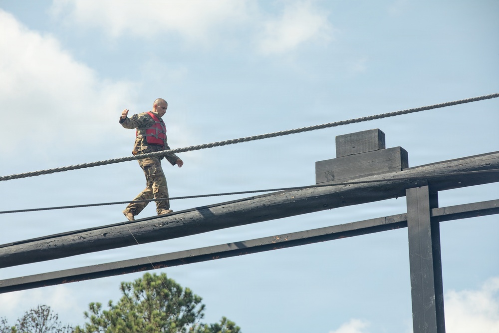 A bald individual wearing military camouflage uniform and a red safety vest is walking along a narrow elevated wooden beam as part of what appears to be a military obstacle course or confidence course. The person is using outstretched arms for balance while navigating the beam at significant height. Rope guide rails are visible on either side of the beam for safety.