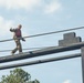 A bald individual wearing military camouflage uniform and a red safety vest is walking along a narrow elevated wooden beam as part of what appears to be a military obstacle course or confidence course. The person is using outstretched arms for balance while navigating the beam at significant height. Rope guide rails are visible on either side of the beam for safety.