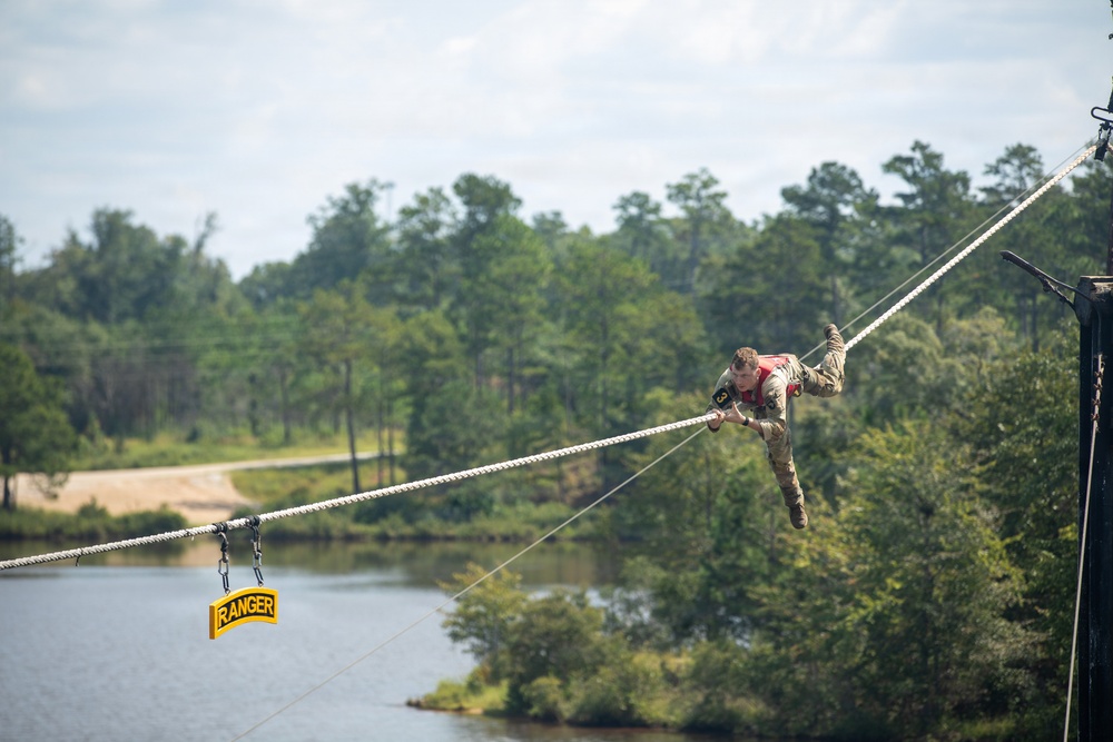 A soldier in military camouflage uniform is traversing a rope obstacle course over a body of water, using both hands and feet to move along the ropes. A yellow 'RANGER' tab sign is visible hanging from one of the lower ropes, indicating this is likely a U.S. Army Ranger training exercise. The setting is an outdoor military training facility surrounded by trees and a lake or pond.