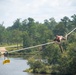 A soldier in military camouflage uniform is traversing a rope obstacle course over a body of water, using both hands and feet to move along the ropes. A yellow 'RANGER' tab sign is visible hanging from one of the lower ropes, indicating this is likely a U.S. Army Ranger training exercise. The setting is an outdoor military training facility surrounded by trees and a lake or pond.