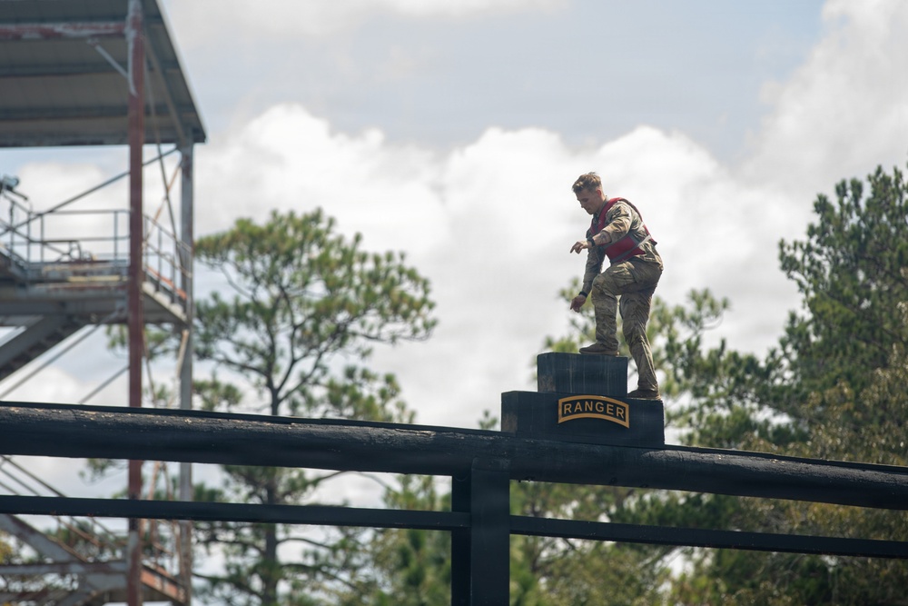 A soldier in camouflage uniform and red safety vest stands atop an elevated obstacle course platform marked with a 'RANGER' tab insignia. The individual appears to be navigating or preparing to traverse a high-elevation training structure. The setting is consistent with a U.S. Army Ranger School or similar military obstacle course training facility.
