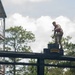 A soldier in camouflage uniform and red safety vest stands atop an elevated obstacle course platform marked with a 'RANGER' tab insignia. The individual appears to be navigating or preparing to traverse a high-elevation training structure. The setting is consistent with a U.S. Army Ranger School or similar military obstacle course training facility.