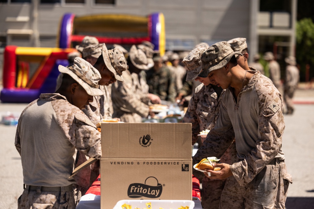 Military personnel in desert camouflage uniforms are gathered outdoors for what appears to be a casual cookout or morale event. Soldiers are serving themselves food from a table with a visible Frito-Lay Vending &amp; Foodservice cardboard box in the foreground. A colorful inflatable bounce house is visible in the background, suggesting a recreational or unit morale event.