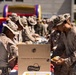 Military personnel in desert camouflage uniforms are gathered outdoors for what appears to be a casual cookout or morale event. Soldiers are serving themselves food from a table with a visible Frito-Lay Vending &amp; Foodservice cardboard box in the foreground. A colorful inflatable bounce house is visible in the background, suggesting a recreational or unit morale event.
