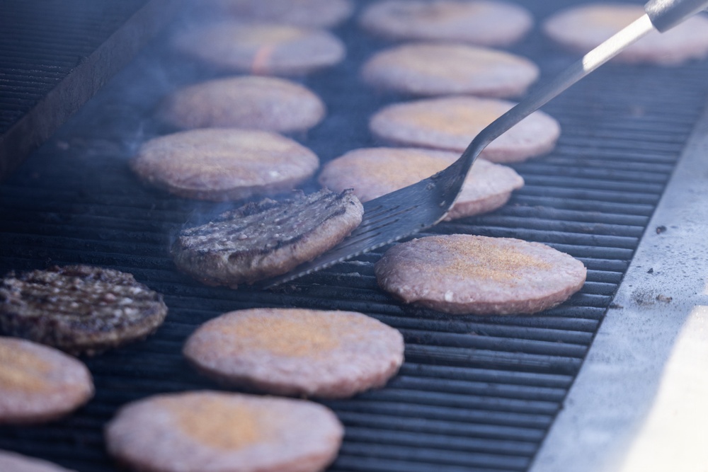 A large commercial grill is loaded with numerous burger patties at various stages of cooking. A metal spatula is actively flipping one patty, which shows visible grill marks and browning on its cooked side. Smoke rises from the grill surface, indicating high heat cooking in what appears to be a large-scale food service setting.
