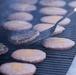 A large commercial grill is loaded with numerous burger patties at various stages of cooking. A metal spatula is actively flipping one patty, which shows visible grill marks and browning on its cooked side. Smoke rises from the grill surface, indicating high heat cooking in what appears to be a large-scale food service setting.