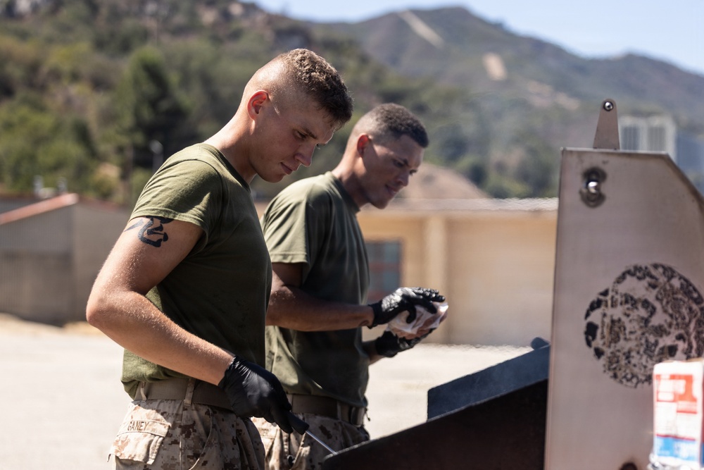 Two military personnel in olive green t-shirts and desert camouflage trousers are performing maintenance or cleaning work outdoors. They are both wearing black gloves and appear to be working on or near a metal structure, likely a shooting target or range equipment. The setting appears to be a military installation with hills and mountains visible in the background.