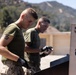 Two military personnel in olive green t-shirts and desert camouflage trousers are performing maintenance or cleaning work outdoors. They are both wearing black gloves and appear to be working on or near a metal structure, likely a shooting target or range equipment. The setting appears to be a military installation with hills and mountains visible in the background.