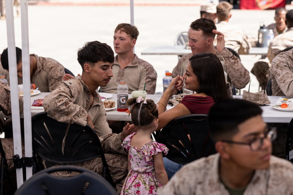 A group of military personnel in desert camouflage uniforms are seated at white tables during what appears to be a social gathering or family day event. A young Marine in the foreground is interacting with a woman in a maroon top and a small child in a floral dress, suggesting a family reunion or unit social event. Multiple service members are visible in the background, some with food and beverages on the tables.