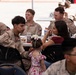 A group of military personnel in desert camouflage uniforms are seated at white tables during what appears to be a social gathering or family day event. A young Marine in the foreground is interacting with a woman in a maroon top and a small child in a floral dress, suggesting a family reunion or unit social event. Multiple service members are visible in the background, some with food and beverages on the tables.