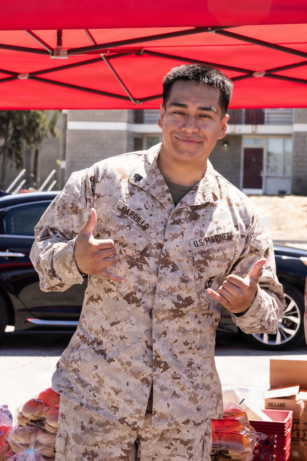 A U.S. Marine in desert MARPAT camouflage uniform is posing with 'shaka' hand gestures under a red pop-up canopy tent. His name tape reads 'CARRILLO' and the branch tape reads 'U.S. MARINES'. The setting appears to be an outdoor community or service event, with food items and supplies visible in the foreground.