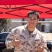 A U.S. Marine in desert MARPAT camouflage uniform is posing with 'shaka' hand gestures under a red pop-up canopy tent. His name tape reads 'CARRILLO' and the branch tape reads 'U.S. MARINES'. The setting appears to be an outdoor community or service event, with food items and supplies visible in the foreground.