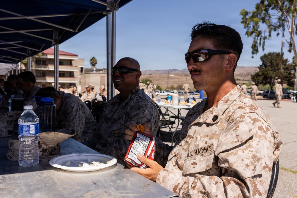 U.S. Marines in desert MARPAT camouflage uniforms are seated at outdoor tables under a blue canopy, eating during what appears to be a field meal or outdoor chow event. The foreground Marine is holding a snack package and has a plate and water bottle on the table. The setting appears to be a military installation with buildings, vehicles, and arid terrain visible in the background.