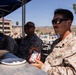 U.S. Marines in desert MARPAT camouflage uniforms are seated at outdoor tables under a blue canopy, eating during what appears to be a field meal or outdoor chow event. The foreground Marine is holding a snack package and has a plate and water bottle on the table. The setting appears to be a military installation with buildings, vehicles, and arid terrain visible in the background.