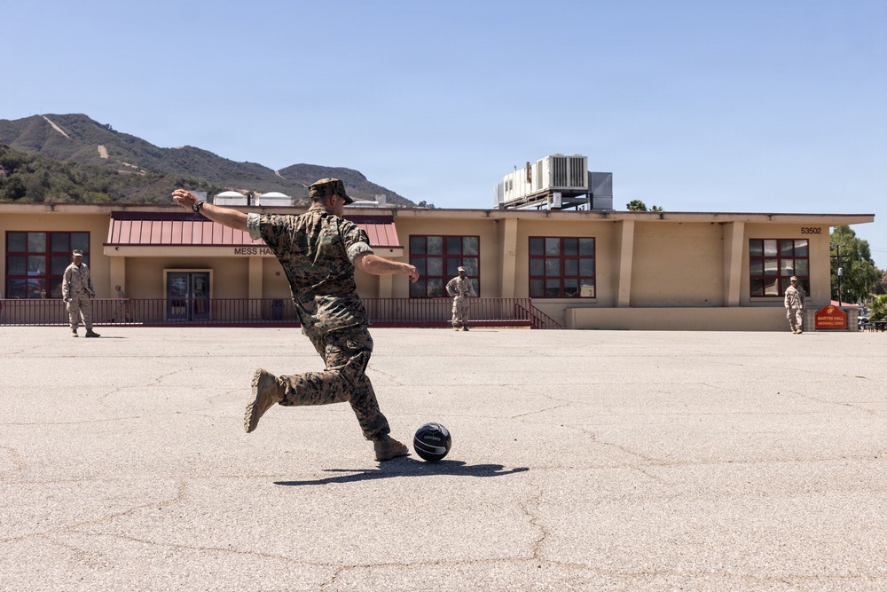 A military service member in camouflage uniform is kicking a ball in an open paved area in front of a building labeled 'Mess Hall' on what appears to be a military base. Several other uniformed personnel are visible in the background near the building. The setting includes mountainous terrain in the background under clear blue skies.