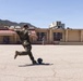 A military service member in camouflage uniform is kicking a ball in an open paved area in front of a building labeled 'Mess Hall' on what appears to be a military base. Several other uniformed personnel are visible in the background near the building. The setting includes mountainous terrain in the background under clear blue skies.