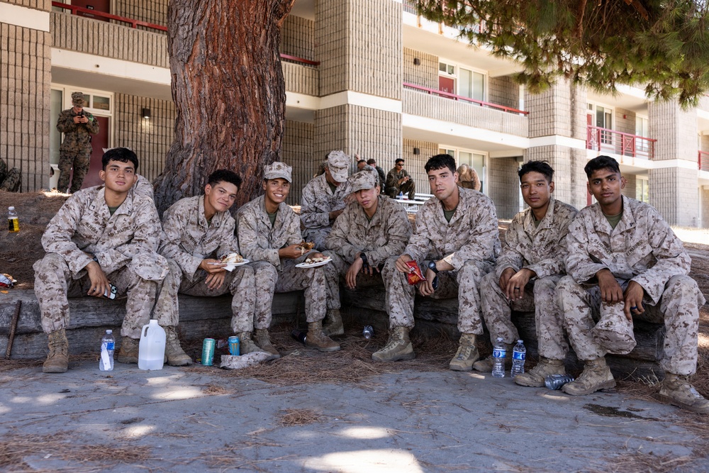 A group of seven young military personnel in MARPAT desert camouflage uniforms are seated together on a low retaining wall beneath a large tree, appearing to be on a break. They are in a relaxed, informal setting outside a multi-story building that resembles military barracks. Several are holding food plates and beverages, suggesting a casual outdoor meal or social gathering.