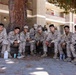 A group of seven young military personnel in MARPAT desert camouflage uniforms are seated together on a low retaining wall beneath a large tree, appearing to be on a break. They are in a relaxed, informal setting outside a multi-story building that resembles military barracks. Several are holding food plates and beverages, suggesting a casual outdoor meal or social gathering.