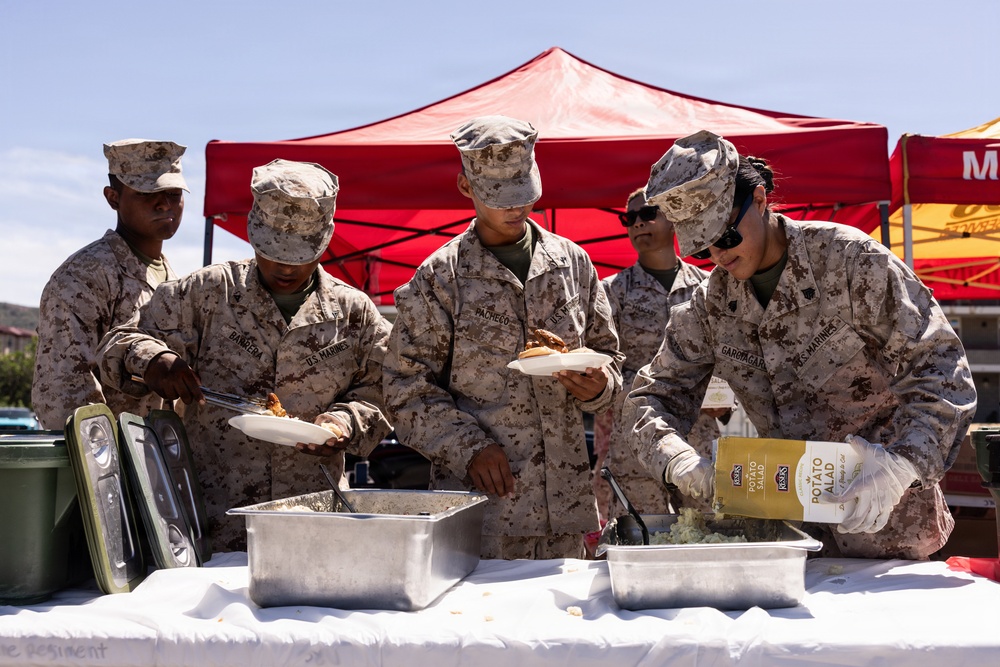 A group of U.S. Marines in desert camouflage uniforms are serving food at an outdoor event, likely a barbecue or unit gathering. Several personnel are working a food service table with metal serving trays, with one Marine pouring potato salad from a store-bought container. A red canopy tent is visible in the background.